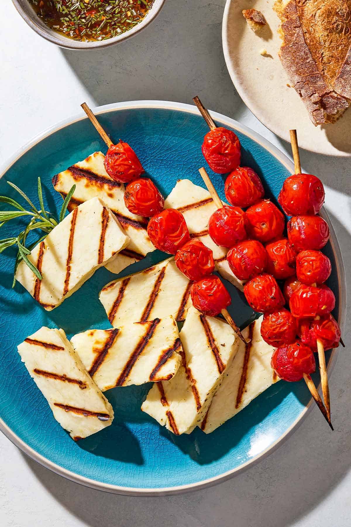 An overhead photo of grilled halloumi with blistered cherry tomatoes, sprigs of rosemary and a small spoon on a serving platter. Next to this is a bowl of the honey-rosemary glaze and a plate with crusty bread.