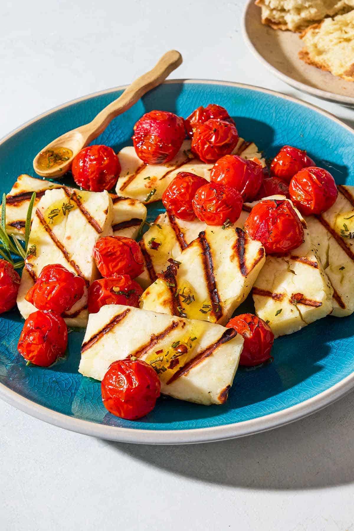 Grilled halloumi with blistered cherry tomatoes, sprigs of rosemary and a small spoon on a serving platter next to a plate with crusty bread.