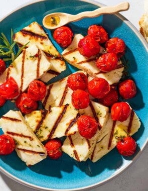 An overhead photo of grilled halloumi with blistered cherry tomatoes, sprigs of rosemary and a small spoon on a serving platter next to some chunks of crusty bread.