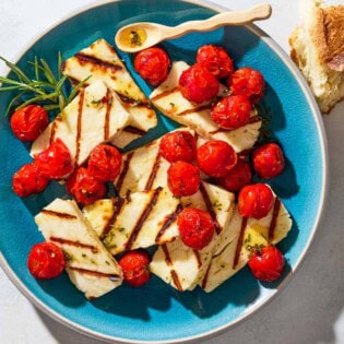 An overhead photo of grilled halloumi with blistered cherry tomatoes, sprigs of rosemary and a small spoon on a serving platter next to some chunks of crusty bread.