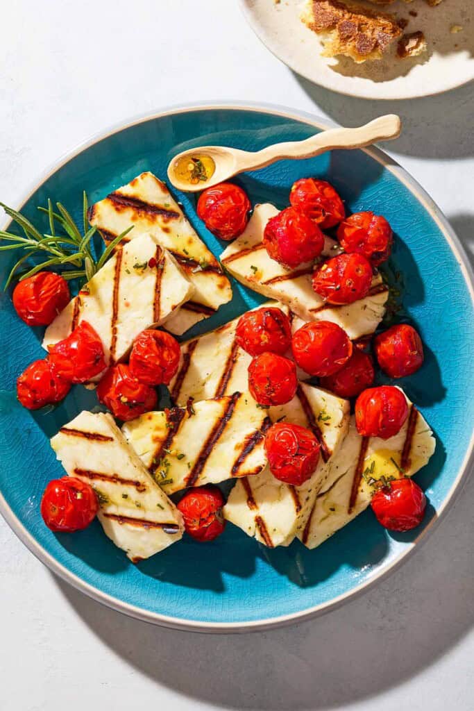 An overhead photo of grilled halloumi with blistered cherry tomatoes, sprigs of rosemary and a small spoon on a serving platter next to a plate with crusty bread.