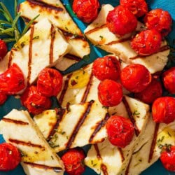 An overhead photo of grilled halloumi with blistered cherry tomatoes, sprigs of rosemary and a small spoon on a serving platter.