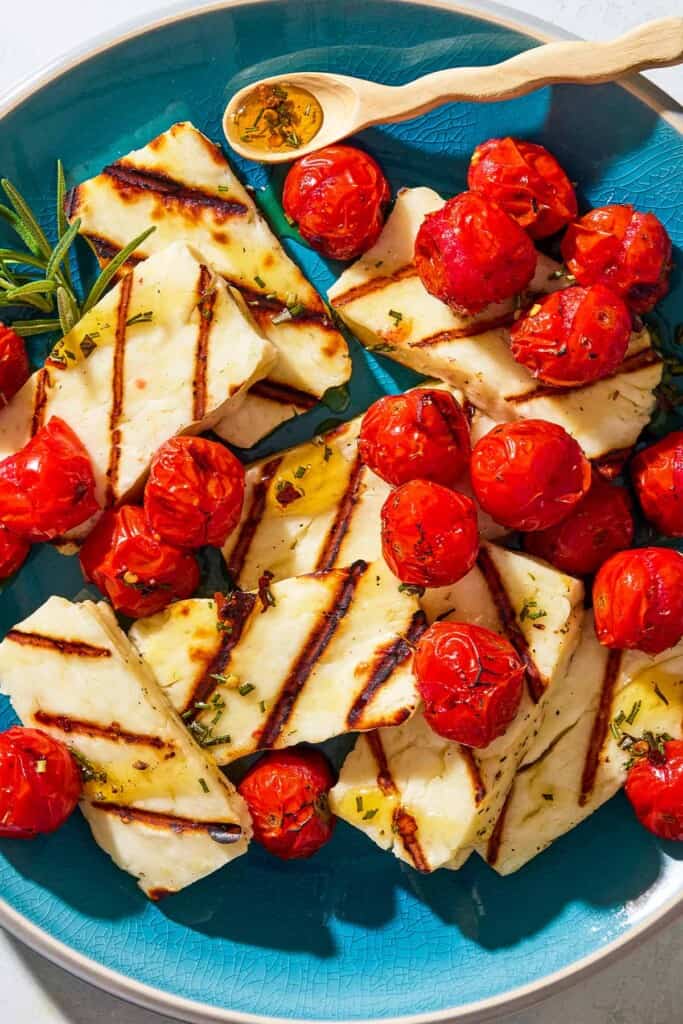 An overhead photo of grilled halloumi with blistered cherry tomatoes, sprigs of rosemary and a small spoon on a serving platter.