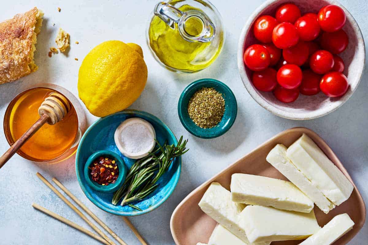 The ingredients for grilled halloumi including halloumi cheese, olive oil, oregano, cherry tomatoes, salt, rosemary, honey, lemon, chili flakes, and some crusty bread.