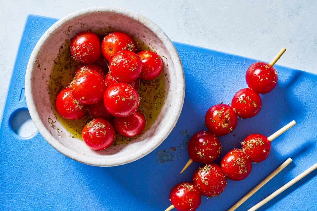 Cherry tomatoes in a bowl with olive oil, oregano and a pinch of salt next to 2 skewers of the tomatoes and two empty skewers sitting on a cutting board.