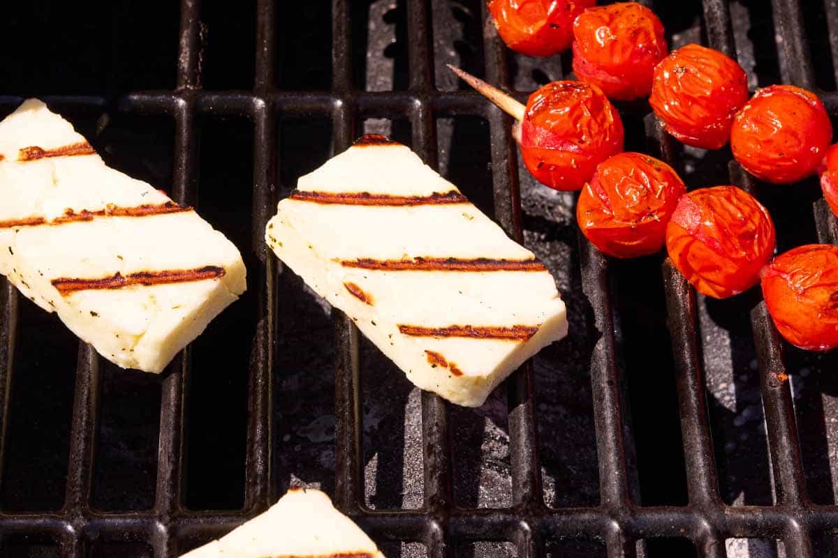 A close up of slices of halloumi cheese and skewers of cherry tomatoes being grilled on an outdoor grill.