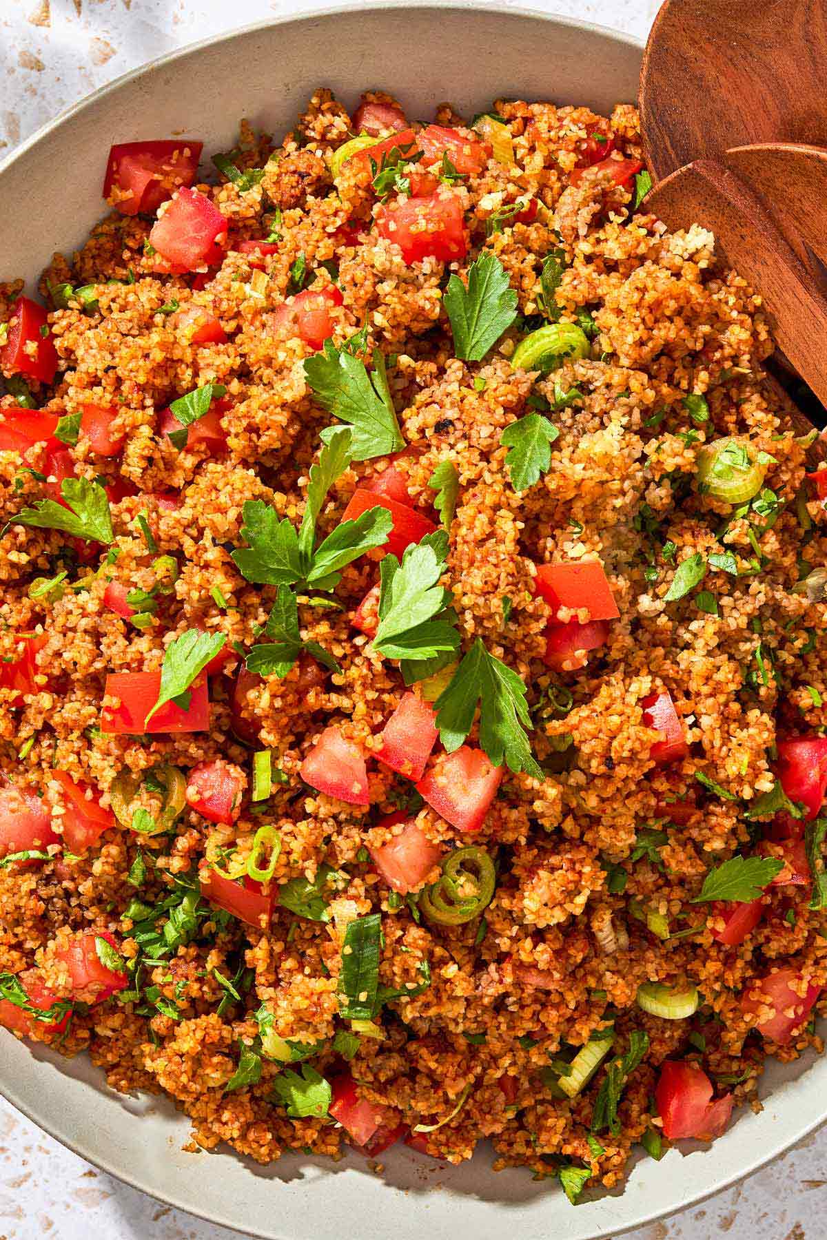 An overhead close up photo of kisir Turkish bulgur salad in a serving bowl with wooden serving utensils.