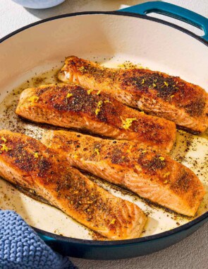 4 salmon filets in a skillet after being pan seared. Next to this is a bowl of spices, a juiced lemon half and a towel.