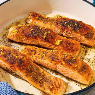 4 salmon filets in a skillet after being pan seared. Next to this is a bowl of spices, a juiced lemon half and a towel.