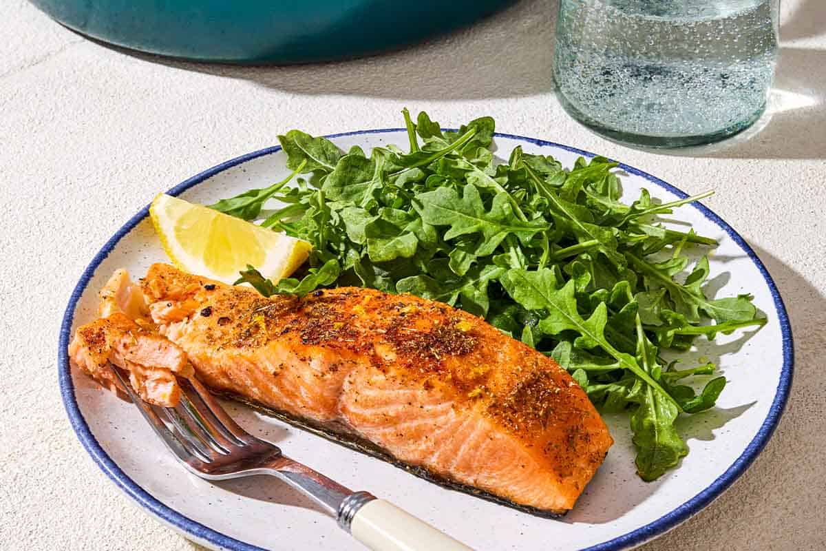 A close up of a pan seared salmon filet on a plate with a fork, lemon wedge, and a side of arugula next to a glass of water.