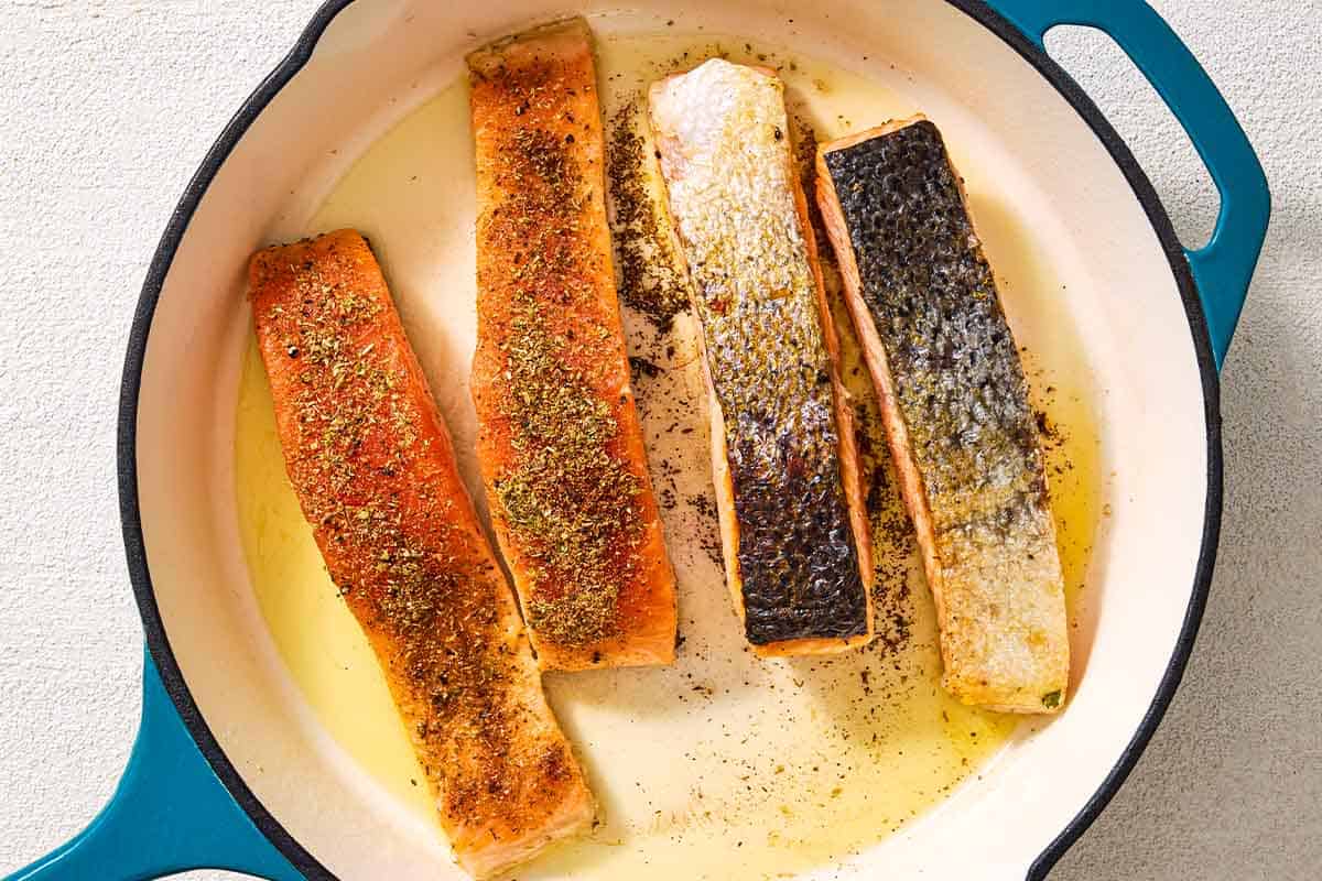 A close up of 4 salmon filets being pan seared in a skillet. Two are flesh side up, and two are skin side up.