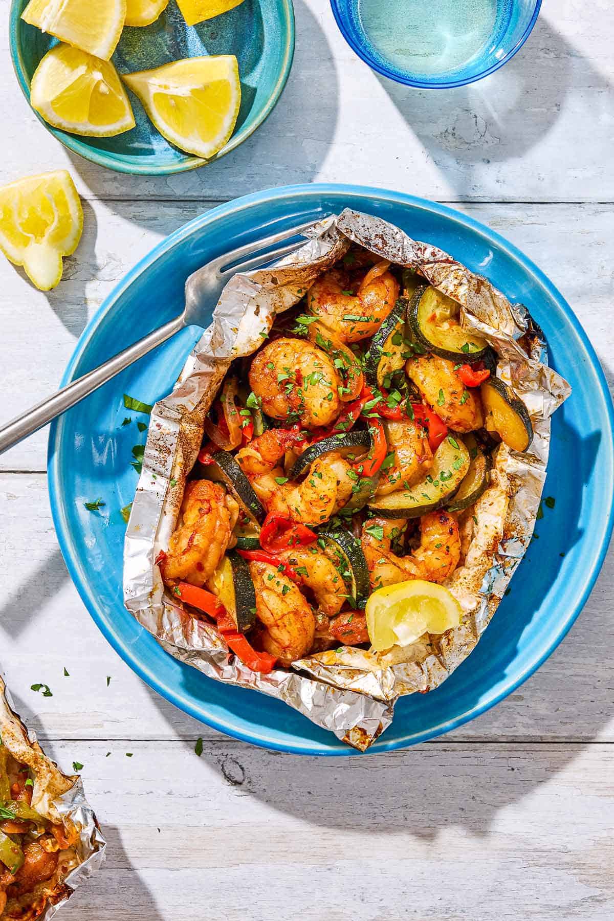 An opened grilled shrimp foil packet with a lemon wedge on a plate with a fork next to a bowl of lemon wedges and a glass of water.