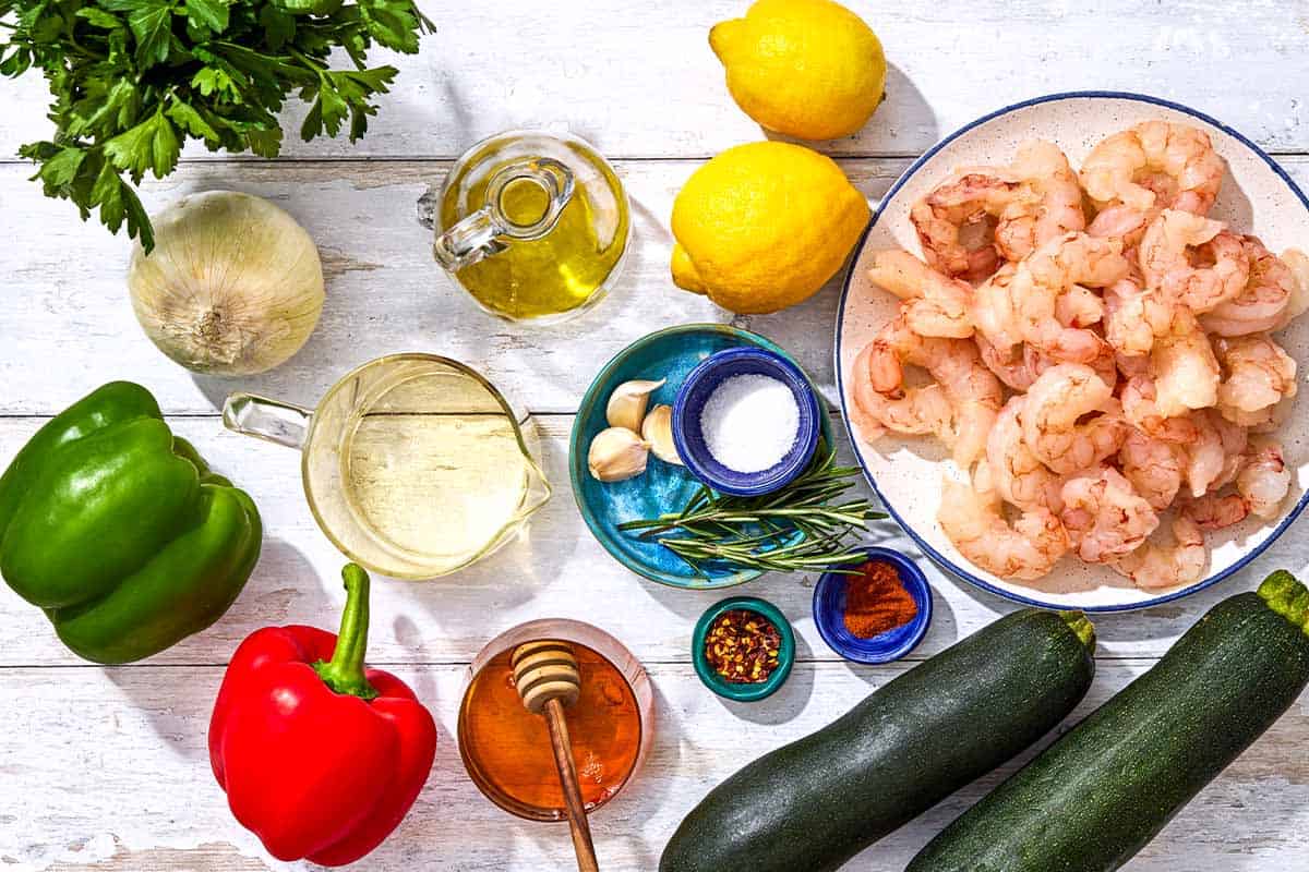 Ingredients for grilled shrimp foil packets including shrimp, zucchini, green bell pepper, red bell pepper, onion, olive oil, white wine, garlic, lemons, smoked paprika, rosemary, chili flakes, honey, salt and parsley.