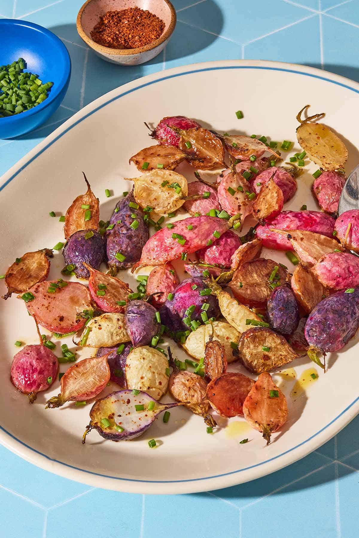 A close up of roasted radishes garnished with chopped chives on a serving platter. Next to this is a bowl of chives, and a bowl of the seasonings.