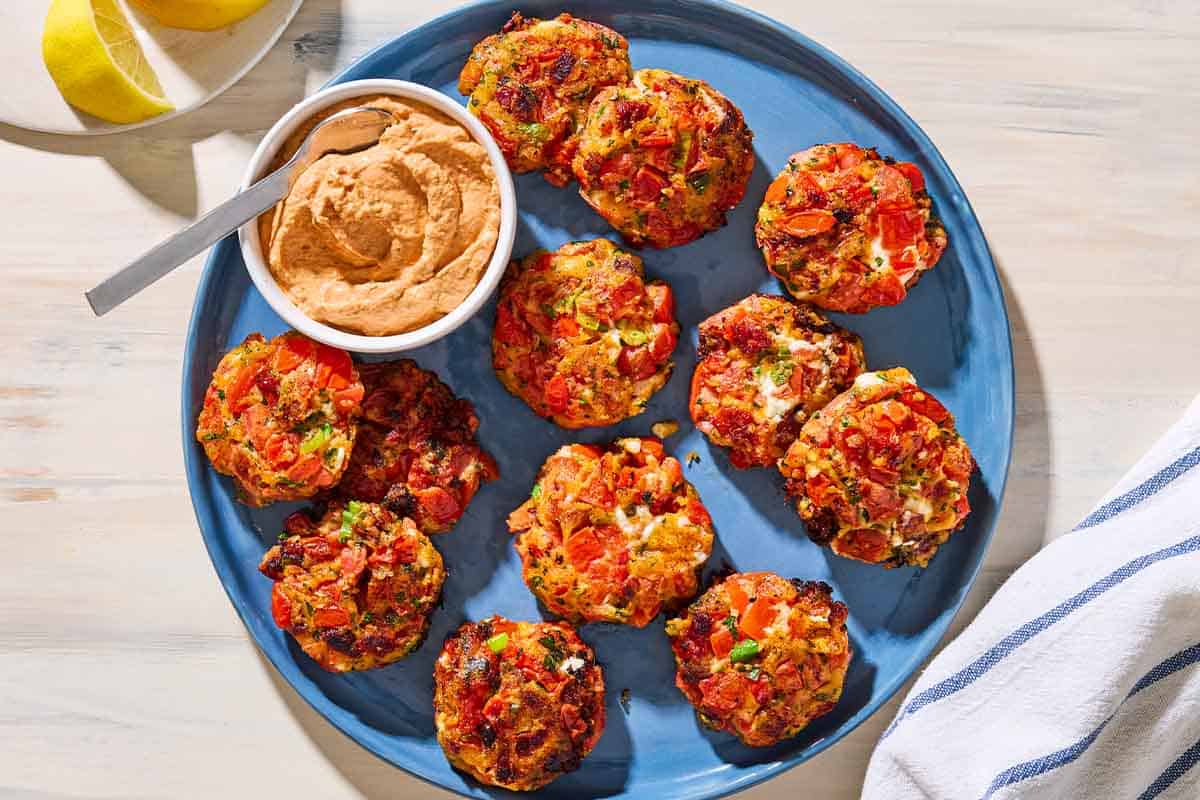 An overhead photo of tomato fritters and a bowl of sun dried tomato yogurt dip with a spoon on a serving platter. Next to this is a plate with lemon wedges and a towel.