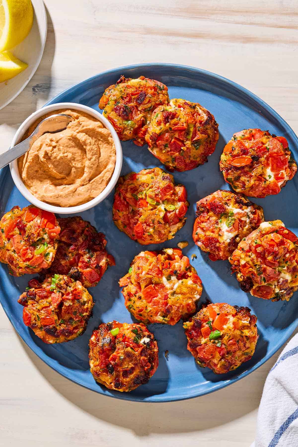 An overhead photo of tomato fritters and a bowl of sun dried tomato yogurt dip with a spoon on a serving platter. Next to this is a plate with lemon wedges and a towel.