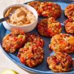 A close up of tomato fritters and a bowl of sun dried tomato yogurt dip with a spoon on a serving platter. Next to this is a plate with lemon wedges and a towel.