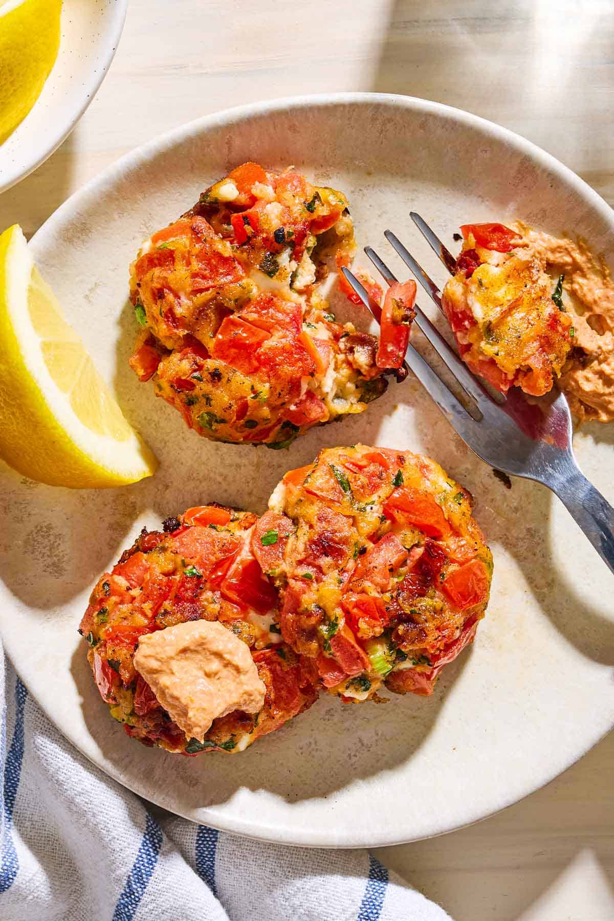 A close up photo of 3 tomato fritters on a plate with a fork and a lemon wedge. One fritter has been topped with a dollop of the dip.