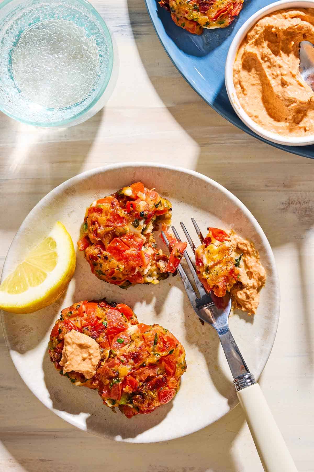 An overhead photo of 3 tomato fritters on a plate with a fork and a lemon wedge. One fritter has been topped with a dollop of the dip. Next to this is a glass of water and the serving plate of fritters and the bowl of dip.