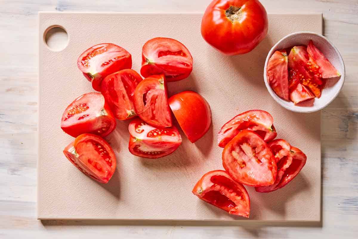 Several tomato wedges on a cutting board, some of them have been seeded. Also on the cutting board is a whole tomato and a bowl with the seeds.