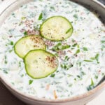 A close up of a bowl of cucumber soup garnished with sumac, parsley and cucumber slices on a plate.