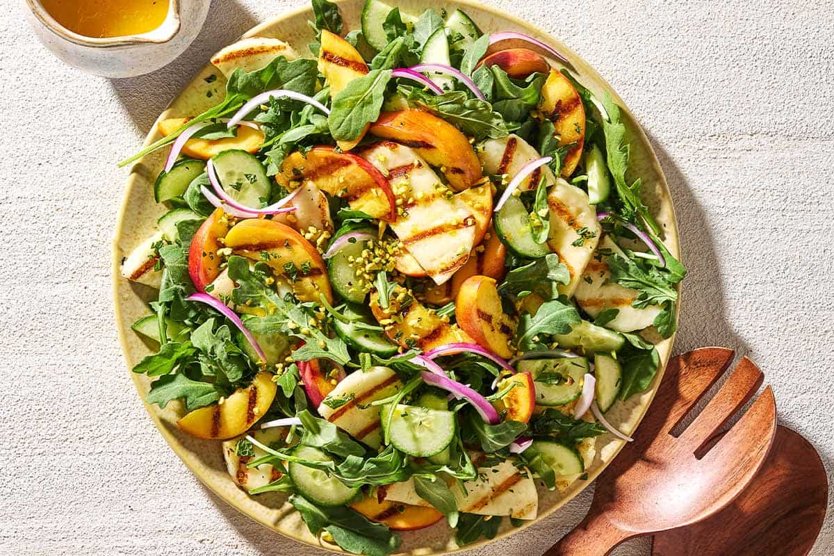 An overhead photo of halloumi and peach salad on a platter next to a small pitcher of dressing and wooden serving utensils.