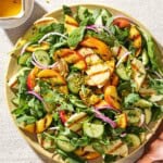 An overhead photo of halloumi and peach salad on a platter next to a small bowl of crushed pistachios, a small pitcher of dressing, and wooden serving utensils.