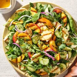 An overhead photo of halloumi and peach salad on a platter next to a small bowl of crushed pistachios, a small pitcher of dressing, and wooden serving utensils.