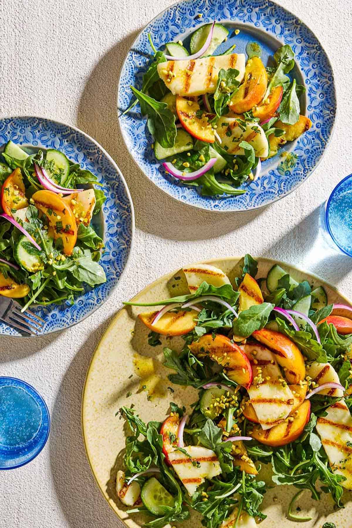 An overhead photo of a two servings of halloumi and peach salad on plates, one with a fork. Next to this is the rest of the salad on a serving platter and 2 glasses of water.