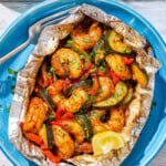 An opened grilled shrimp foil packet with a lemon wedge on a plate with a fork next to a plate of lemon wedges.