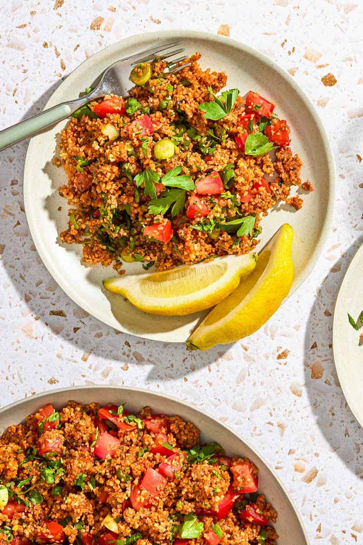 An overhead photo of a serving of kisir Turkish bulgur salad on a plate with a fork and two lemon wedges. Next to this is the rest of the salad in a serving bowl.