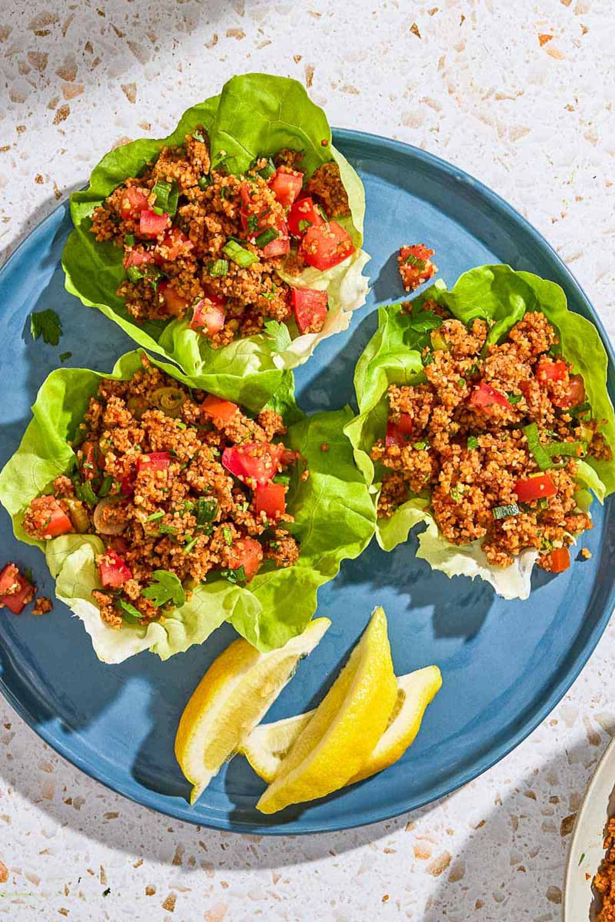 An overhead photo of 3 servings of kisir Turkish bulgur salad in lettuce boats on a plate with three lemon wedges.