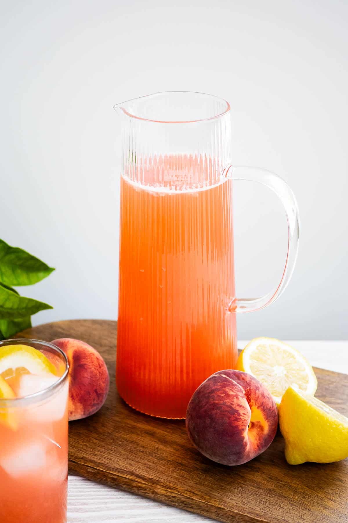 A pitcher if peach lemonade on a wooden tray with peaches and lemons. Next to this is a glass of peach lemonade.