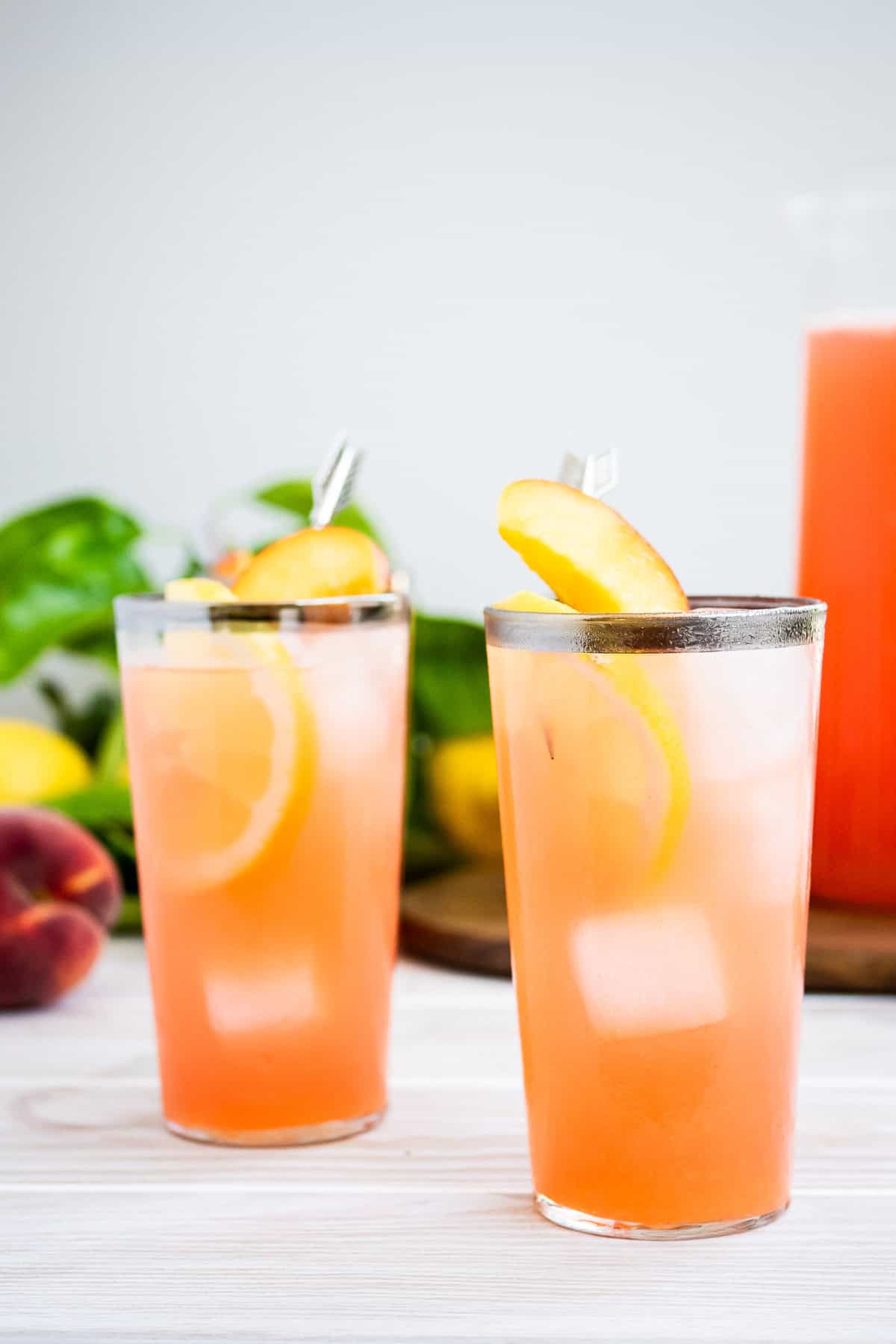 A close up of two glasses of peach lemonade in front of a pitcher of the lemonade on a wooden tray, as well as some peaches and lemons.