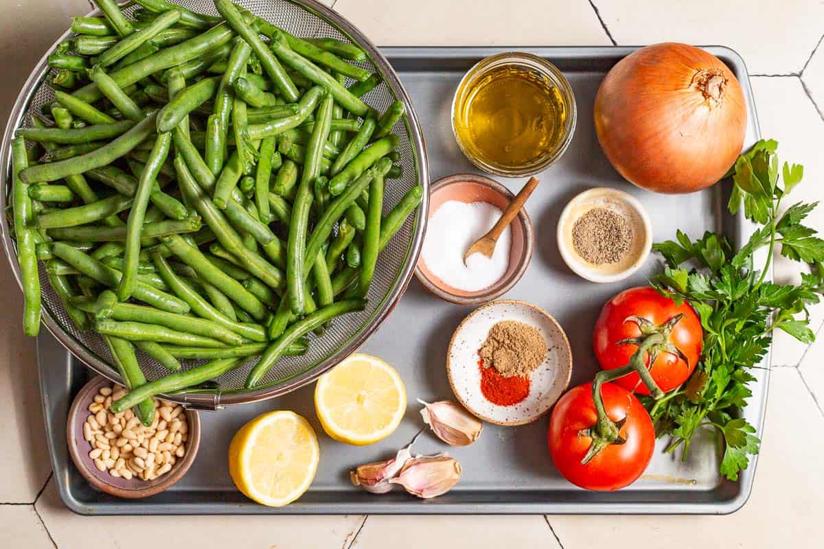 Ingredients for sauteed green beans including green beans, olive oil, onion, salt, garlic, tomatoes, coriander, paprika, black pepper, pine nuts, lemon, and parsley.