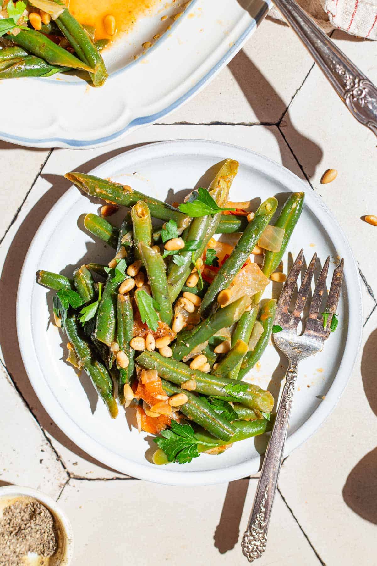 An overhead close up photo of a serving of sauteed green beans on a plate with a fork. Next to this is a serving platter with the rest of the green beans and a bowl of black pepper.