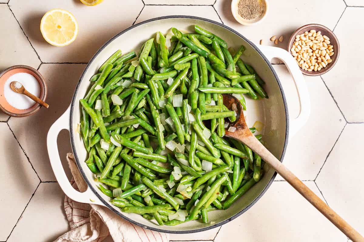 Green beans and chopped onions being sauteed in a skillet with a wooden spoon. Next to this is a kitchen towel, a lemon half and bowls of salt, black pepper and pine nuts.