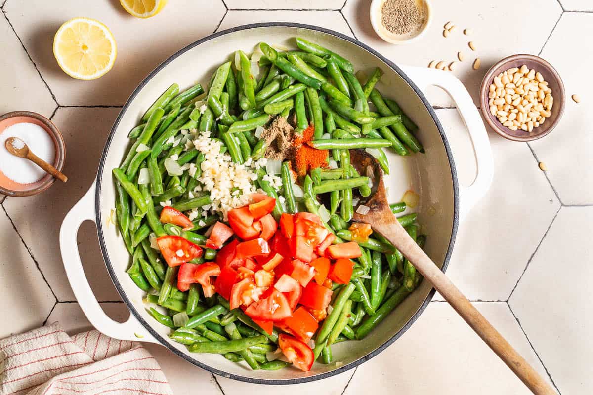 Green beans, onions, garlic, tomatoes and spices being sauteed in a skillet with a wooden spoon. Next to this is a kitchen towel, two lemon halves and bowls of salt, black pepper and pine nuts.