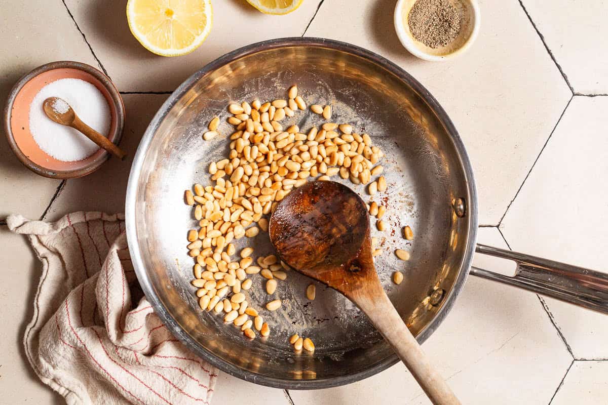 Pine nuts being toasted in a skillet with a wooden spoon. Next to this is a kitchen towel, two lemon halves and bowls of salt, black pepper and pine nuts.