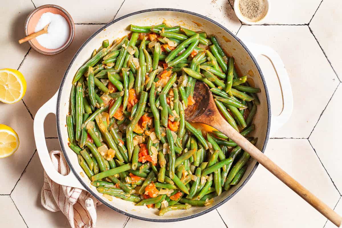 Green beans, onions, garlic, and tomatoes being sauteed in a skillet with a wooden spoon. Next to this is two lemon halves and bowls of salt, black pepper and pine nuts.