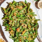 An overhead photo of sauteed green beans on a serving platter with a spoon. Surrounding this is a lemon half, kitchen towel, parsley, and bowls of salt, black pepper and pine nuts.