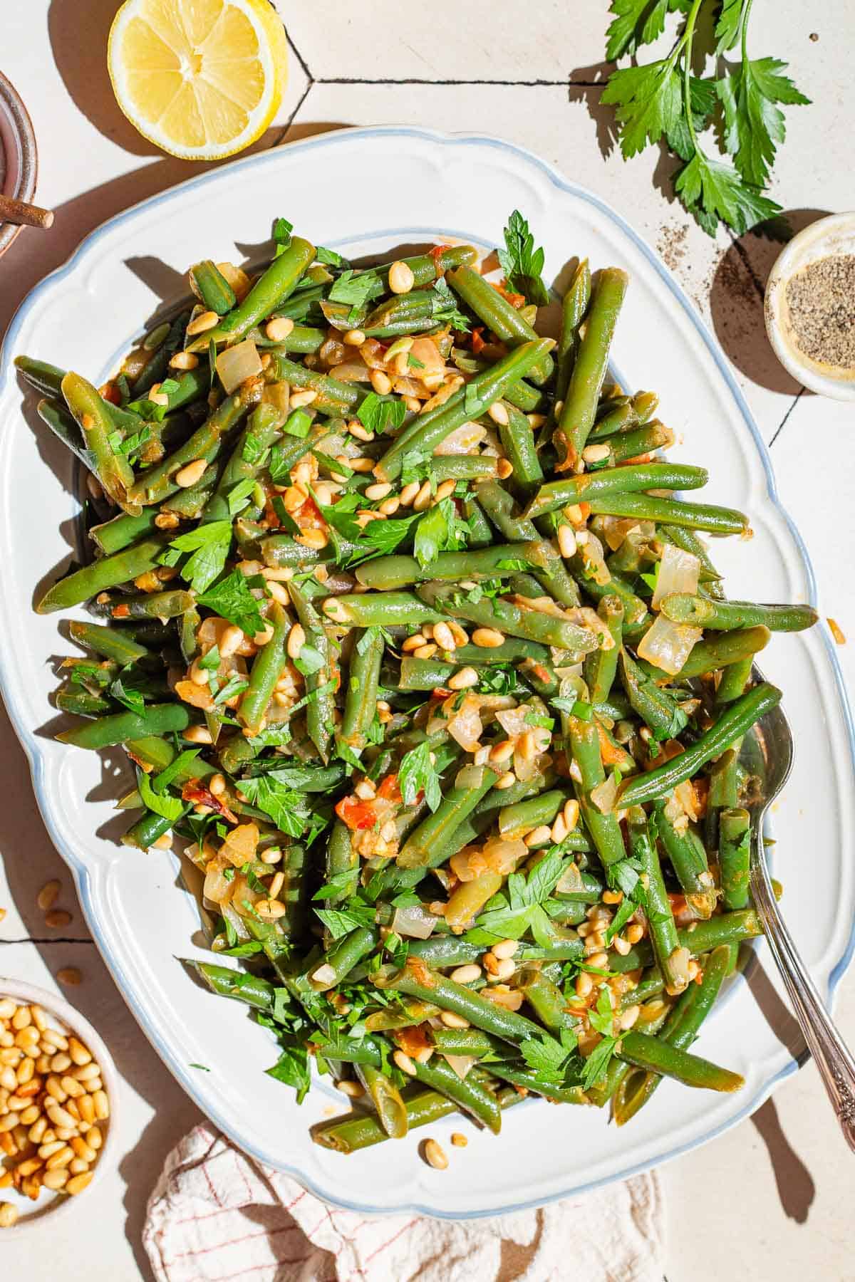 An overhead photo of sauteed green beans on a serving platter with a spoon. Surrounding this is a lemon half, kitchen towel, parsley, and bowls of salt, black pepper and pine nuts.