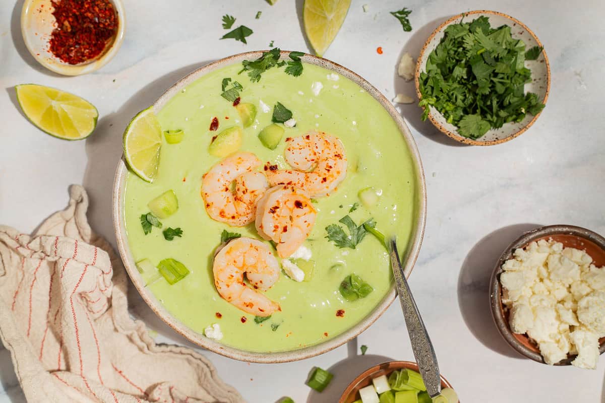 An overhead photo of avocado soup topped with shrimp, cilantro, feta, aleppo pepper and scallions in a bowl with a spoon. Next to this is a kitchen towel, lime wedges, and bowls of aleppo, feta, cilantro and scallions.