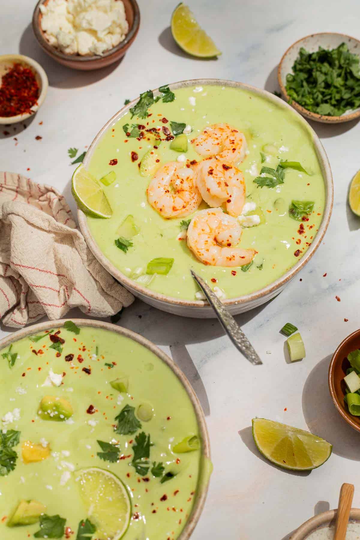 A close up of avocado soup topped with shrimp, cilantro, feta, aleppo pepper and scallions in a bowl with a spoon. Next to this is another bowl of the soup with a spoon, a kitchen towel, lime wedges, and bowls of aleppo, feta, and cilantro.