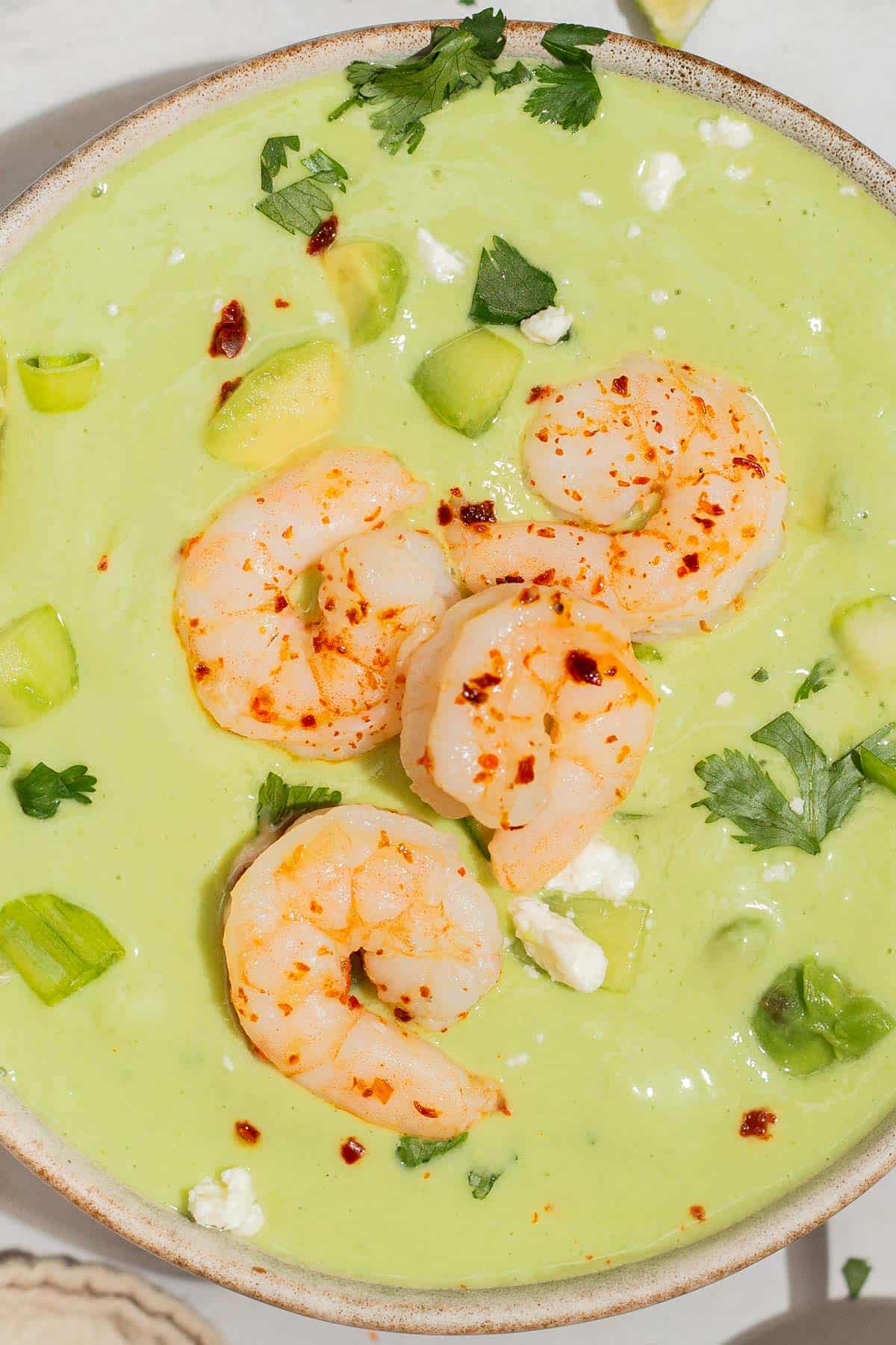 A close up of avocado soup topped with shrimp, cilantro, feta, aleppo pepper and scallions in a bowl.