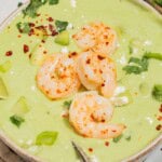 A close up of avocado soup topped with shrimp, cilantro, feta, aleppo pepper and scallions in a bowl with a spoon.