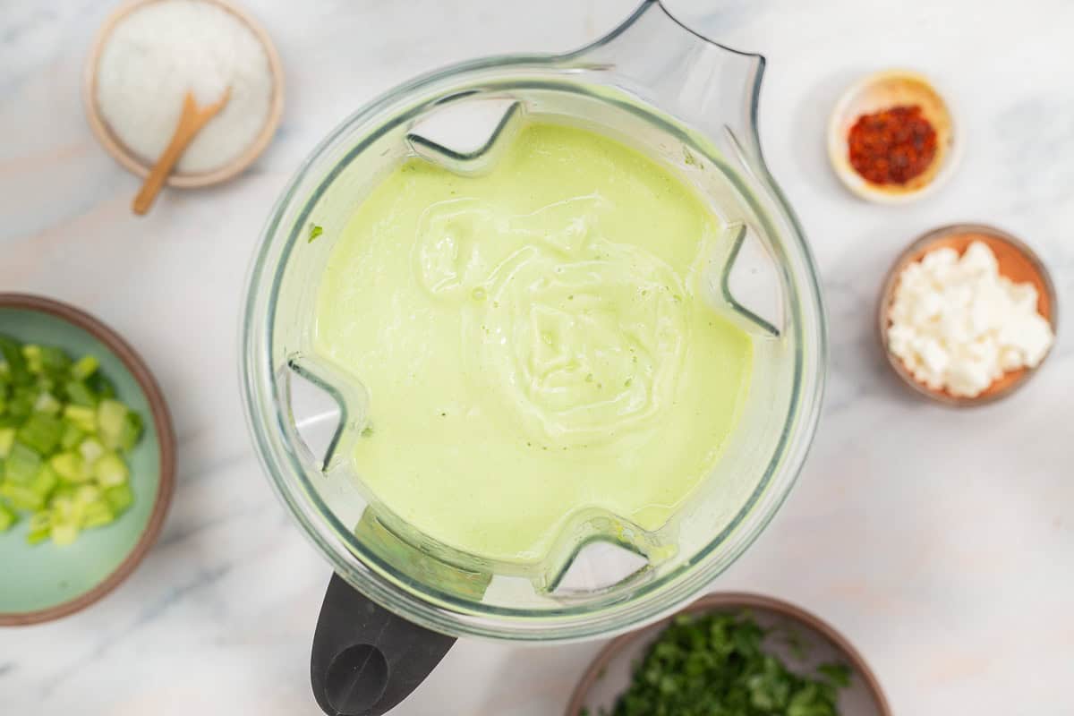 An overhead photo of the ingredients for the avocado soup mixed together in the pitcher of a blender. Next to this are bowls of chopped scallions, salt, aleppo, feta, and cilantro.