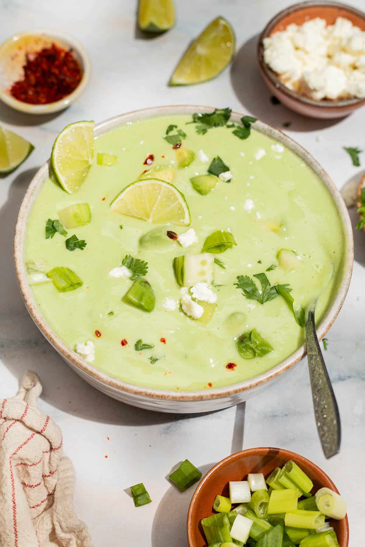 A close up of avocado soup garnished with lime wedges, feta, cilantro, aleppo pepper and scallions in a bowl with a spoon. Next to this is a kitchen towel, lime wedges, and bowls of chopped scallions, parsley and feta cheese.