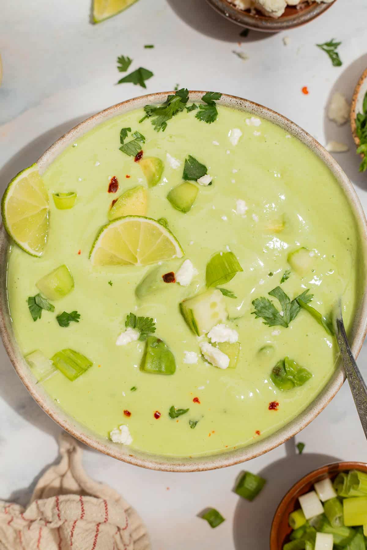 An overhead photo of avocado soup garnished with lime wedges, feta, cilantro, aleppo pepper and scallions in a bowl with a spoon. Next to this is a kitchen towel and a bowl of scallions.