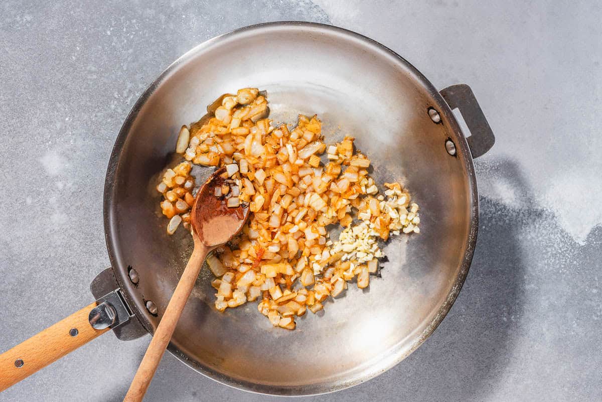 An overhead photo of garlic and onion being sauteed in a skillet with a wooden spoon.